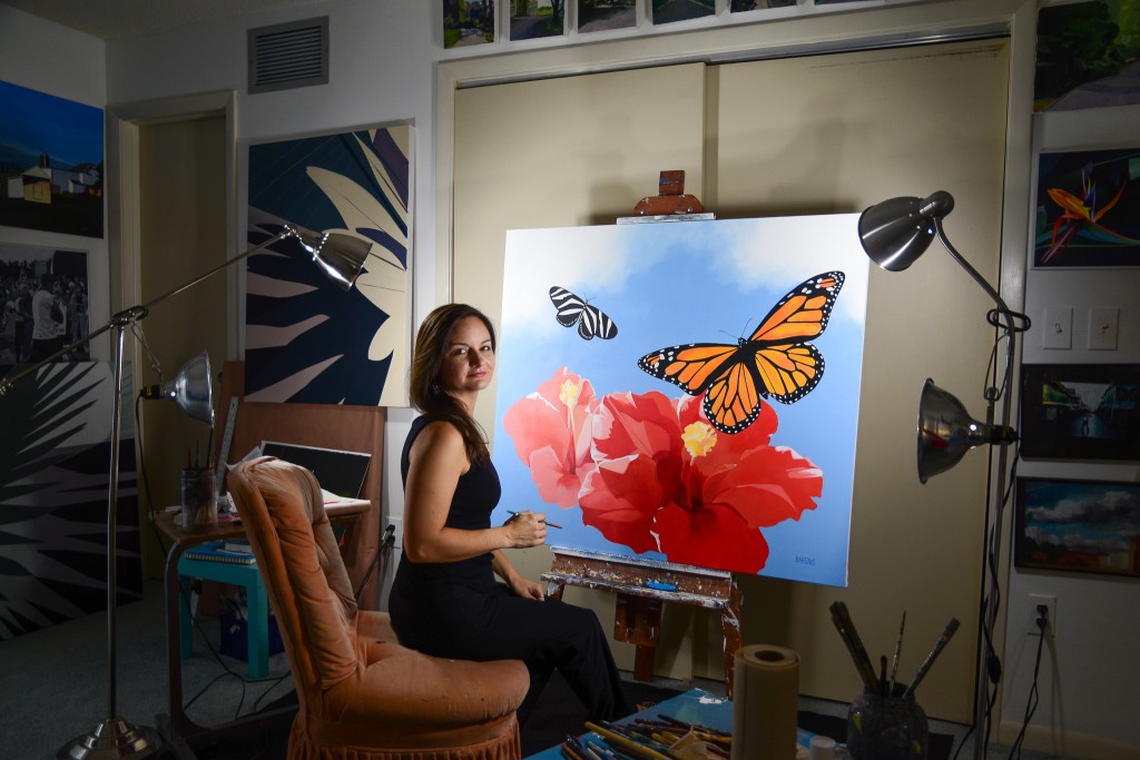 Elizabeth Barenis sits in her studio, in front of her painting of butterflies and hibiscus.