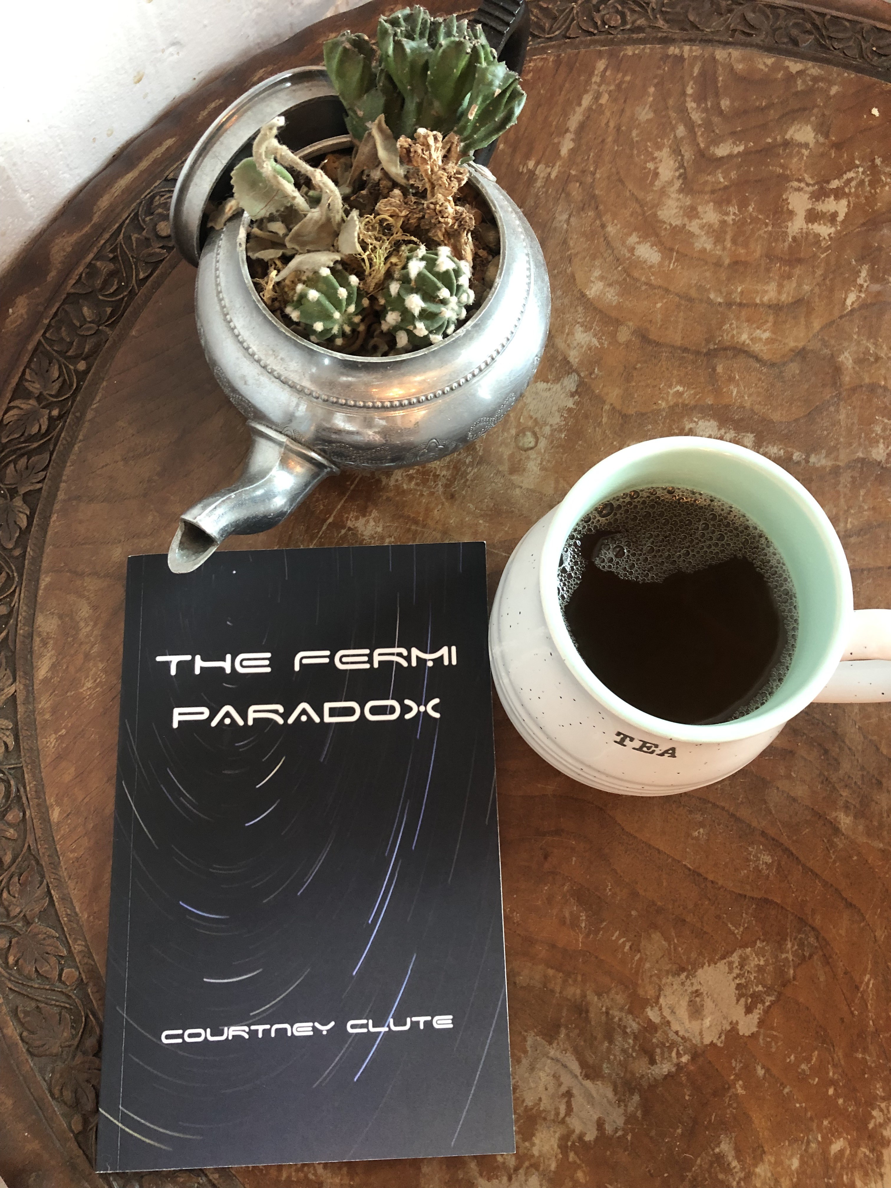 The book "The Fermi Paradox" by Courtney Clute sits flat on a table next to a cup of tea and a potted succulent.